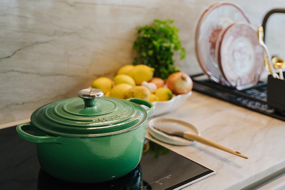 Bancada de cozinha com uma cocotte em verde e suporte para colher na cor creme da Le Creuset