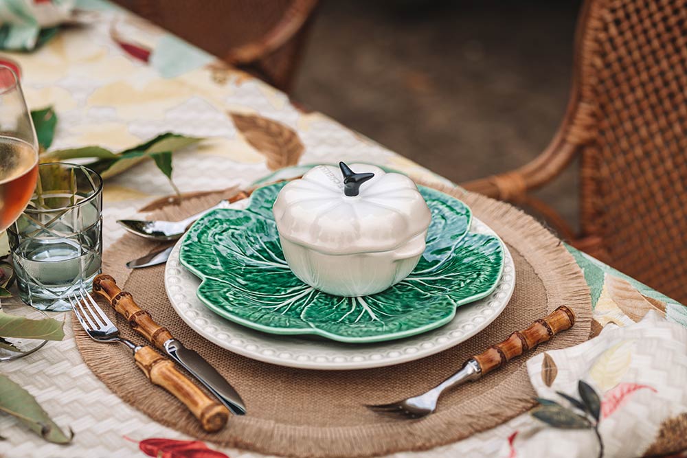 Mesa com toalha de mesa com padrão outonal, individual em rattan, prato raso branco, com prato em forma de couve por cima e cocotte branca da Le Creuset.