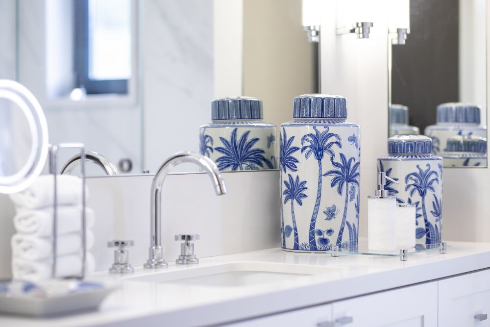 Bancada de casa de banho organizada com potes em porcelana com palmeiras azuis e brancas e toalheiro de bancada cinzento.
