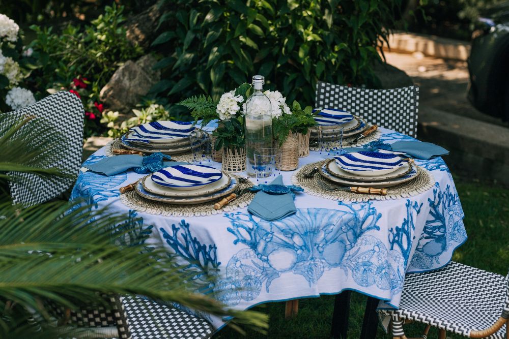 Mesa de jardim cm uma toalha azul e branca que remete para os elementos do mar. A mesa está posta com pratos azuis e brancos, marcadores com búzios, copos com bolas azuis e centro de mesa com flores brancas.